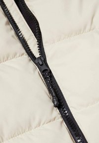 Beige Daunenjacke mit schwarzem Reißverschluss, ausgestattet mit einem strukturierten Reißverschlussanhänger und einem glatten Futter. Die Nähte sind entlang der Nahtstellen sichtbar.