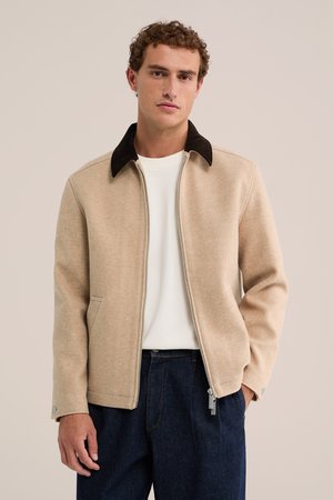 Jeune homme portant une veste zippée beige à col noir, chemise blanche et jean bleu foncé, se tenant devant un fond clair uni.