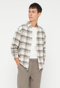 Πουκάμισο φλιάνελ σε καρώ σχέδιο με κρέμα, μαύρο και καφέ χρώμα, διαθέτει γιακά με κουμπιά και μακριά μανίκια, σε συνδυασμό με γκρι παντελόνια φόρμας με κορδόνια.