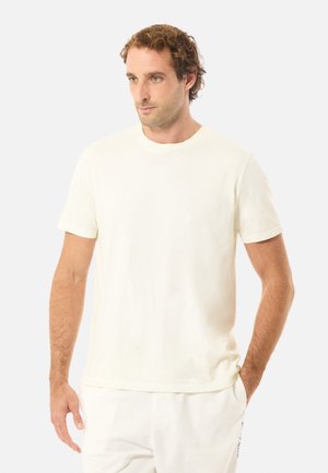 Mies, jolla on vaaleanruskeat kiharat hiukset, pukeutunut yksinkertaiseen valkoiseen lyhythihaiseen t-paitaan ja valkoisiin housuihin, seisoo yhdellä kädellä taskussa.