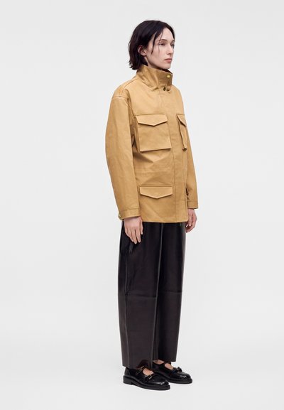 Personne portant une veste beige avec de grandes poches frontales, un pantalon noir à jambes larges et des chaussures noires, debout devant un fond clair uni.