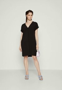 Mulher com cabelo trançado, vestindo um vestido preto com decote em V, sandálias azuis de salto aberto, colar de conchas e segurando uma pequena bolsa lilás clara.