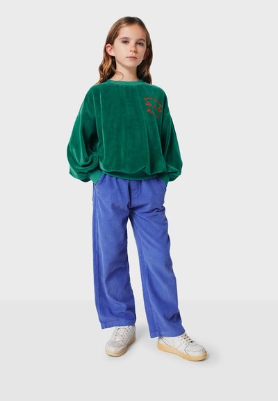 Groen fluwelen sweatshirt met rode bloemenborduursels, gecombineerd met losse blauwe corduroybroeken en witte sneakers. Ontspannen pasvorm.