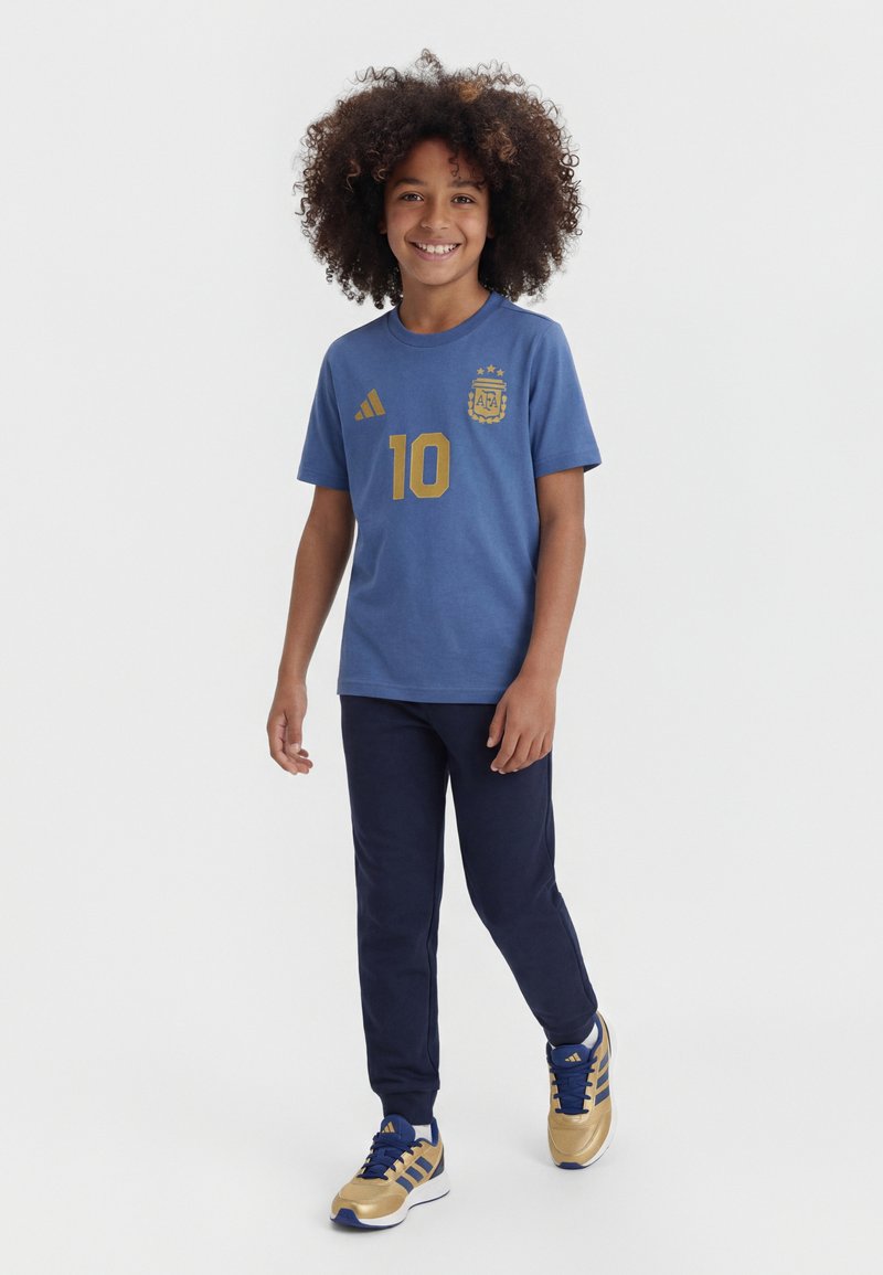 Παιδί με σγουρά μαλλιά φορώντας μια μπλε φανέλα ποδοσφαίρου της Αργεντινής, μπλε σκούρο παντελόνι και χρυσά-μπλε αθλητικά παπούτσια Adidas, στέκεται και χαμογελάει.