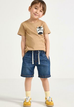 Ung dreng smiler, iført en beige t-shirt med et zebra motiv, denim shorts med snore, gule sneakers og lyse sokker.