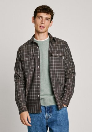 Flannel shirt in dark brown with white grid pattern and two chest pockets, layered over a light green knit sweater. Denim jeans complete the outfit.