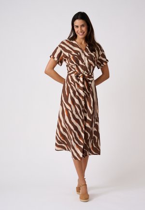 Femme souriante portant une robe midi imprimé zèbre marron, crème et orange avec boutons et ceinture à la taille, assortie à des sandales compensées couleur beige.
