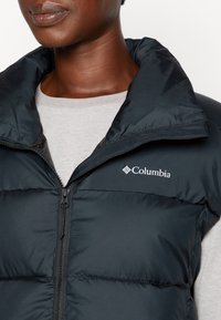 Juodas paminkštintas liemenė su aukštu apykakle ir užtrauktuku. Viršuje kairėje pusėje yra "Columbia" logotipas. Dėvima ant šviesiai pilkos marškinėlių.