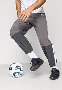 Γκρι αθλητικά παντελόνια με ελαστικές μανσέτες, πλευρικές ρίγες και μια διακριτική λογότυπο. Μαύρα ποδοσφαιρικά παπούτσια και λευκές κάλτσες. Μία μπάλα ποδοσφαίρου στο πόδι.