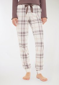 Donna che indossa pantaloni da pigiameria bianchi a quadri con linee viola e rosa e una maglietta a maniche lunghe color malva, in piedi a piedi nudi su uno sfondo bianco.