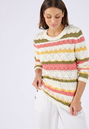 Femme portant un pull en tricot crème avec des rayures horizontales jaunes, roses et vertes, tenant l'ourlet, assorti à un pantalon blanc sur fond uni.