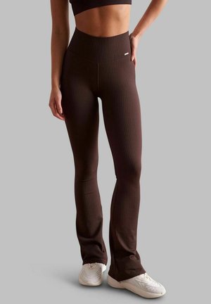 Femme portant des leggings évasés côtelés marron foncé taille haute et des baskets blanches, debout avec une main sur la hanche sur fond gris.