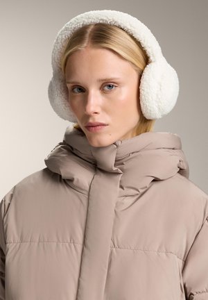 Cagule din blană artificială albă, formă rotundă cu textură pufoasă. Modelul poartă o jachetă bej cu umplutură, cu guler înalt și detalii de cusătură.