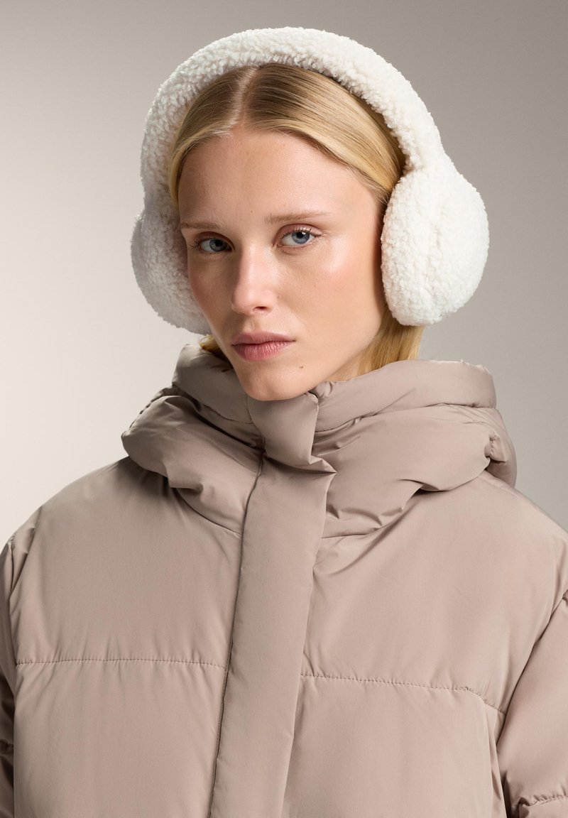 Cuffie in finta pelliccia bianca, di forma rotonda con texture soffice. Il modello indossa una giacca imbottita beige con colletto alto e dettagli delle cuciture.