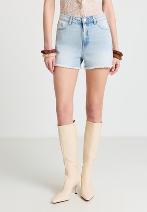 Donna che indossa pantaloncini di denim azzurro sfilacciato, stivali al ginocchio beige con tacco alto, top beige in pizzo e braccialetti spessi su entrambi i polsi.