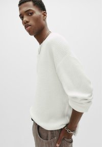 Junger Mann, der einen weißen strukturierten Pullover und braune Hose trägt, mit einer silbernen Kette und einem Armband, posiert vor einem schlichten Hintergrund.