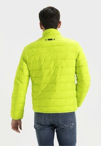 Lys neon grøn pufferjakke med horisontalt quiltet mønster, høj krave og et lille mærketag på bagsiden. Båret med blå jeans.