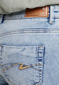 Jeans in denim azzurro chiaro con etichetta in pelle, caratterizzati da una tasca posteriore con un motivo ricamato e dettagli di cucitura in vista.