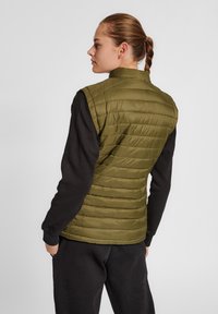 Olivgrön pufferväst med kviltat mönster och hög krage, bärs över en svart långärmad skjorta. Slätt, lätt material.