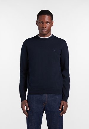 Uomo con maglione blu scuro semplice e jeans blu, in piedi davanti a uno sfondo bianco.