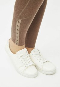 Zapatillas blancas con superficie texturizada y suela plana, combinadas con leggings marrones que presentan botones de presión en el lateral y un diseño ajustado.