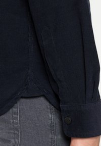 Laivastonsininen puuvillahousupaita, jossa on napillinen kalvosin, tikatut saumat ja kärkikaarikaulus; kankaan pinta on ribbineulosta ja pehmeä.