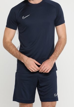 Homme portant un t-shirt de sport Nike bleu foncé et un short, debout les mains jointes devant un fond uni.
