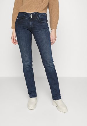 Person iført mørkeblå slim fit jeans, beige strikket sweater og hvide chunky støvler, der står på en ensartet hvid baggrund.