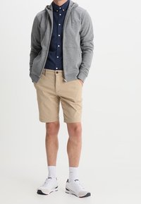 Grijze zip-up hoodie over een donkerblauw overhemd met knopen, gecombineerd met kaki korte broek en witte sneakers met rode en marineblauwe accenten.