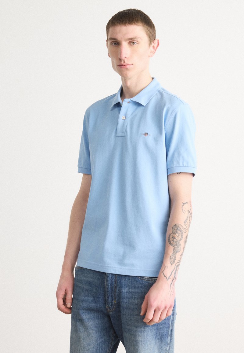 Lyseblå polo skjorte laget av bomull, med tre-knapps placket og korte ermer. Den har en liten brodert logo på brystet.