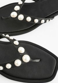 Infradito nere con suola piatta, dotate di un cinturino a forma di V decorato con perle finte bianche rotonde e dettagli in tonalità argento.