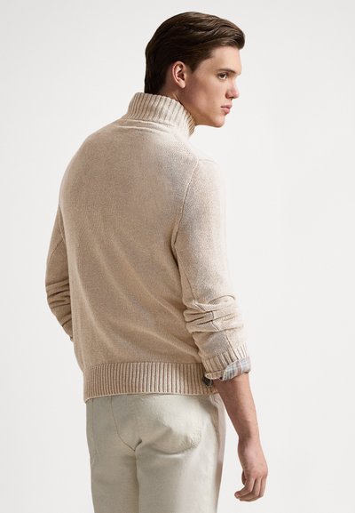 Beige gebreide trui met een hoge hals, geribbelde manchetten en zoom, en opgerolde mouwen die een geruit patroon eronder onthullen.