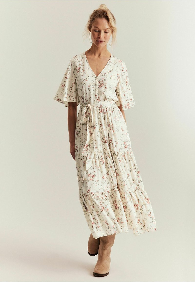 Frau mit blondem Haar, die ein weißes Midikleid mit Blumenmuster und flatternden Ärmeln sowie beige Knöchelstiefel trägt, steht vor einem schlichten Hintergrund.