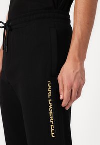 Schwarze Jogginghose mit einem goldenen Logo an der Seite, elastischem Bund und Kordelzug. Der Stoff sieht glatt aus und hat sichtbare Seitentaschen.