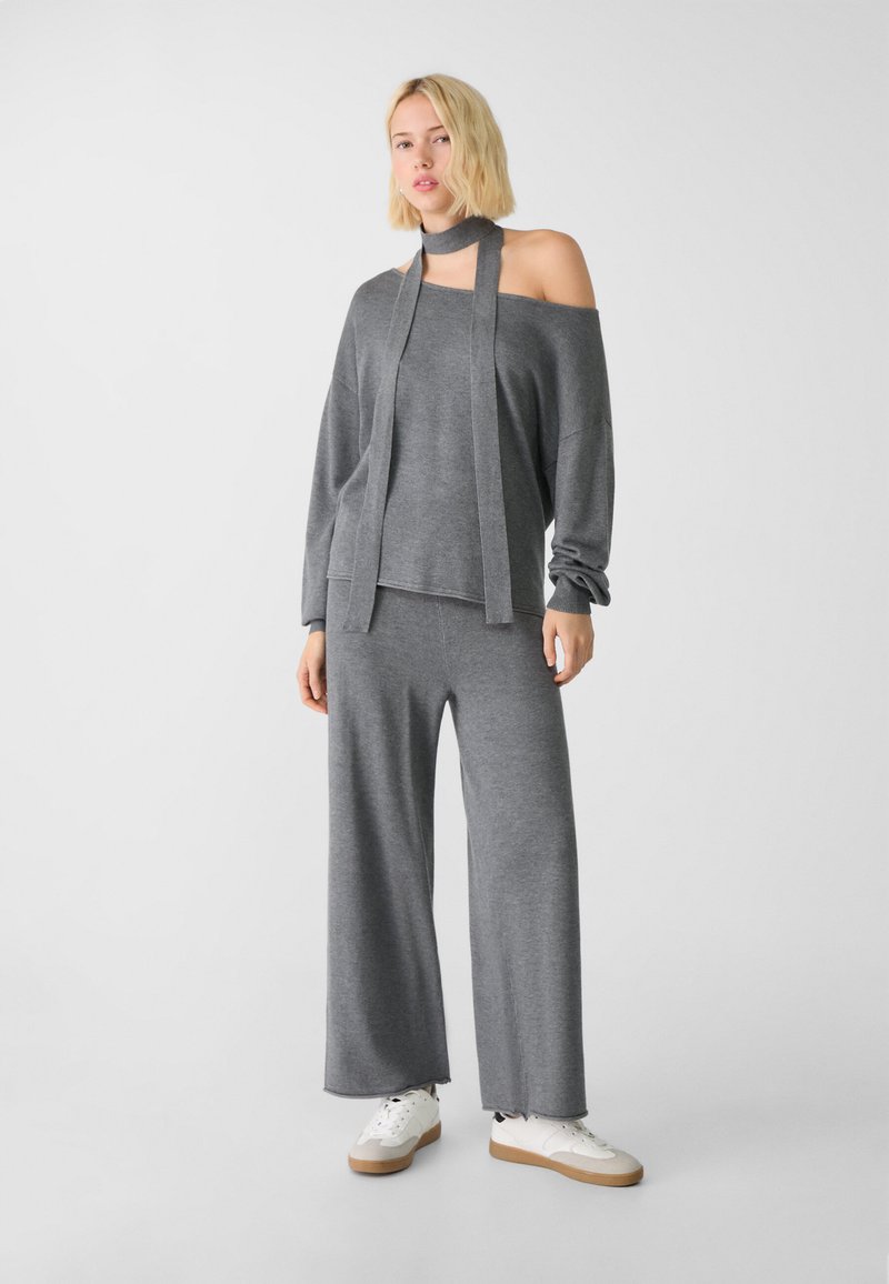Szary komplet dresowy z plecionki, składający się z bluzki na jedno ramię z przymocowanym szalem oraz szerokich spodni. Miękka faktura, luźny krój, w połączeniu z białymi trampkami.