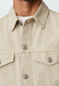 Chaqueta de mezclilla beige con cierre de botones en la parte delantera, cuello puntiagudo y dos bolsillos en el pecho. Presenta botones plateados y una tela texturizada.