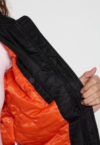 Czarna pikowana kurtka z pomarańczową satynową podszewką, z zapięciem na zatrzaski i gładką wewnętrzną fakturą, ukazująca wyraźny kontrast kolorów.