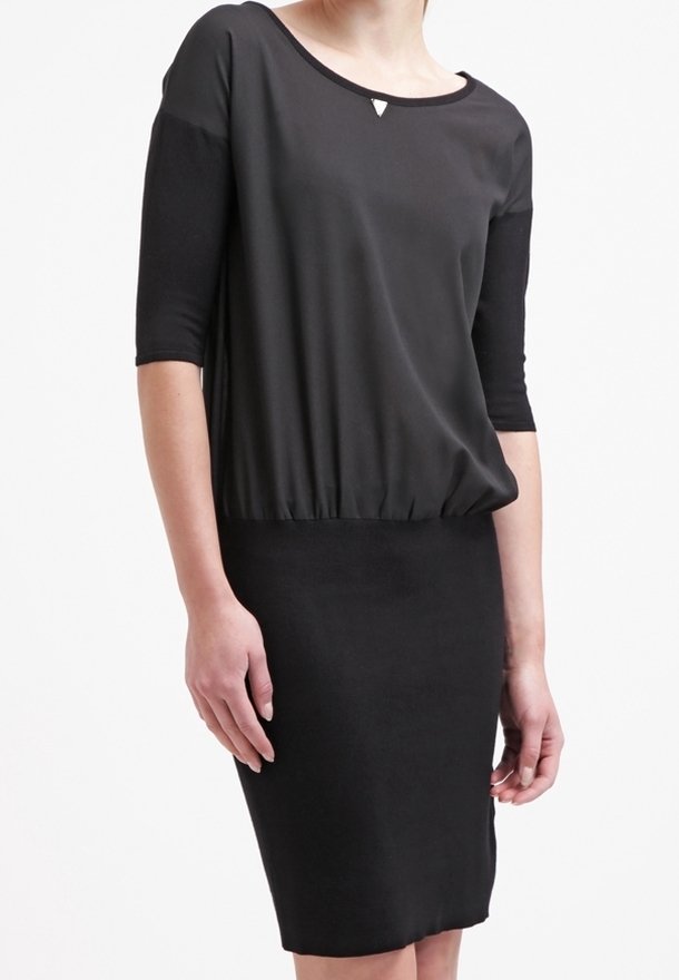 Schwarzes Kleid mit lockerem Oberteil und tailliertem Rock. Hat Dreiviertelärmel, einen runden Halsausschnitt und ein kleines dreieckiges Detail auf der Brust.