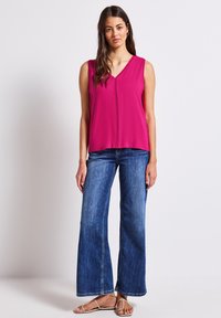Top senza maniche fucsia con scollatura a V, caratterizzato da un dettaglio con cucitura centrale, abbinato a jeans blu a gamba larga e sandali di colore chiaro.
