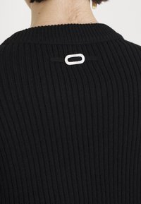 Black ribbed sweater featuring a round neckline and a silver rectangular hardware detail at the back. Textured fabric with vertical lines.