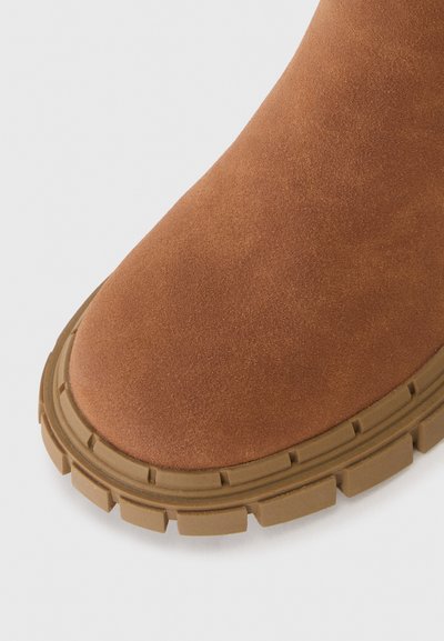 Bota de tornozelo em camurça castanha com bico arredondado, apresentando uma sola de borracha grossa e texturizada. A sola possui padrões de aderência visíveis para tração.