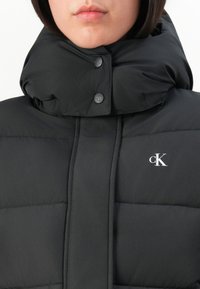 Personne portant une doudoune noire avec un col montant et un logo "cK" visible sur la poitrine, les boutons fermés au niveau du col.