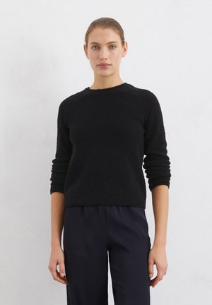 Schwarzer, strukturiert gestrickter Pullover mit Rundhalsausschnitt und langen, umgeschlagenen Ärmeln. Kombiniert mit dunklen, locker sitzenden Hosen.