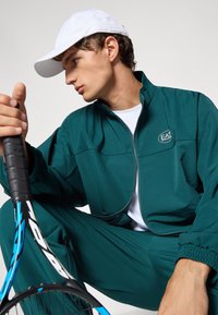 Mladý muž v tyrkysovej teplákovej súprave a bielej šiltovke sedí s modro-čiernou tenisovou raketou v ruke a pozerá sa do strany na jednoliatom pozadí.