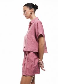 Camicetta corta a maniche corte e pantaloncini coordinati in morbido tessuto rosa chiaro con una finitura strutturata. Presenta dettagli con bottoni e una vestibilità rilassata.