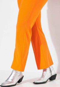 Pantaloni attillati di un vivace arancione con una texture liscia, abbinati a stivaletti neri lucidi con punta leggermente appuntita e tacco a blocco.