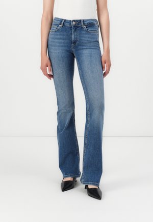 Kobieta w niebieskich prostych dżinsach i czarnych czółenkach z ostrym noskiem, stojąca na jednolitym białym tle.