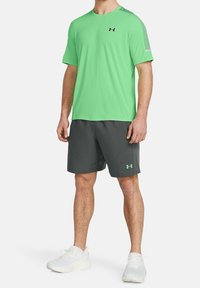 Zelené sportovní tričko s krátkým rukávem a kulatým výstřihem, doplněné tmavě šedými šortkami. Obě položky nesou logo Under Armour. Bílé sportovní boty.