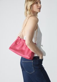 Borsa in pelle rosa con parte superiore arricciata, dotata di una tracolla in catena dorata. La borsa è tenuta di lato contro una maglietta bianca e jeans blu.
