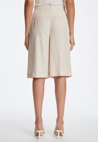 Beigeweite Shorts mit einer glatten Textur und einem knielangen Schnitt, mit einem diskreten Reißverschluss hinten und einem sauberen Abschluss am Saum.